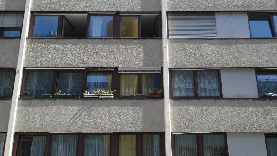 Close-up facade of a concrete apartment building with multiple windows, balconies, curtains and potted plants in muted grey and warm wood tones.