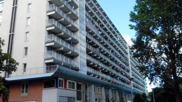 Tall modern apartment building with rows of balconies and soft greenery in the foreground under a clear daylight sky.
