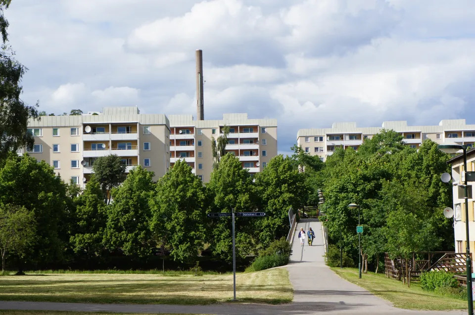Photograph of residential apartment blocks with balconies, mature green trees and a pathway under a soft grey-blue sky, evoking a European urban neighbourhood.